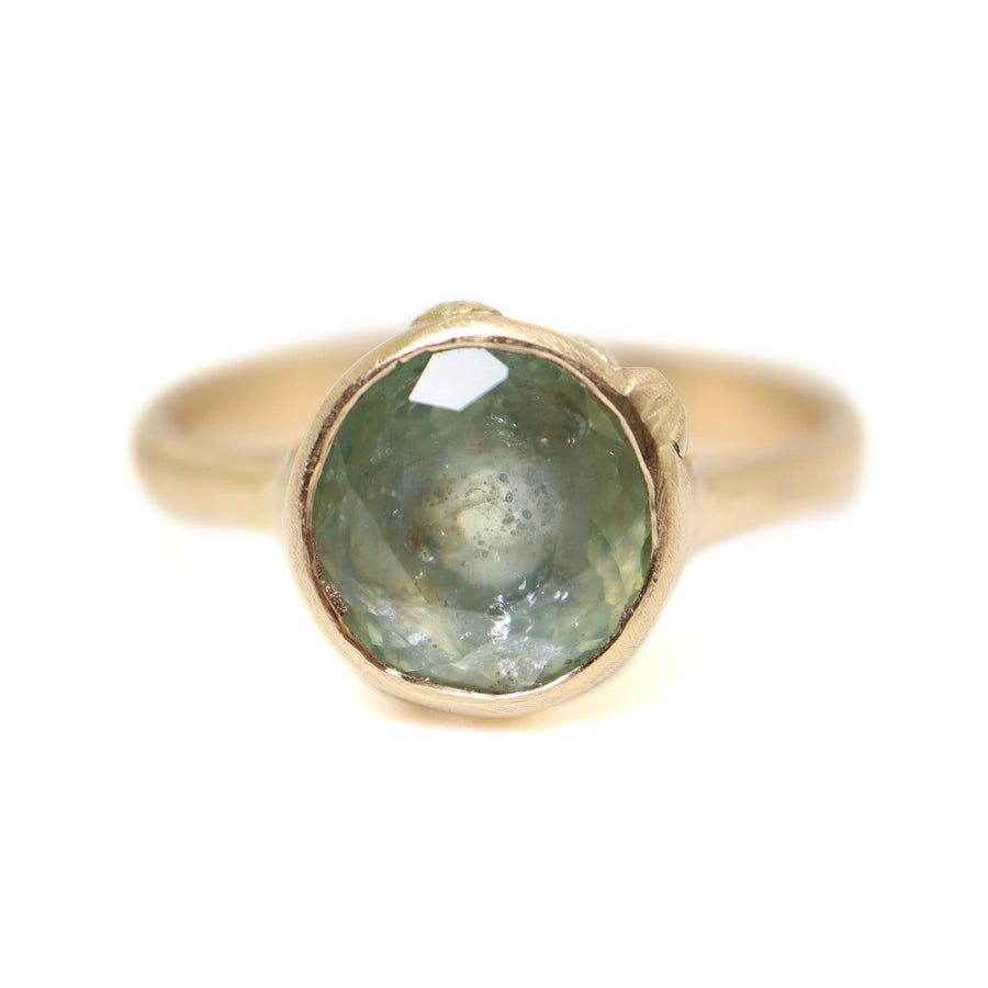 A 2.29ct Montana Sapphire ring. The Montana Sapphire is a pale, murky blue with tinges of green. The Montana sapphire is set in 14k gold with a tapered 14k gold band. The setting is encrusted with 14k gold barnacles. The ring is viewed off center and sits on a white background.