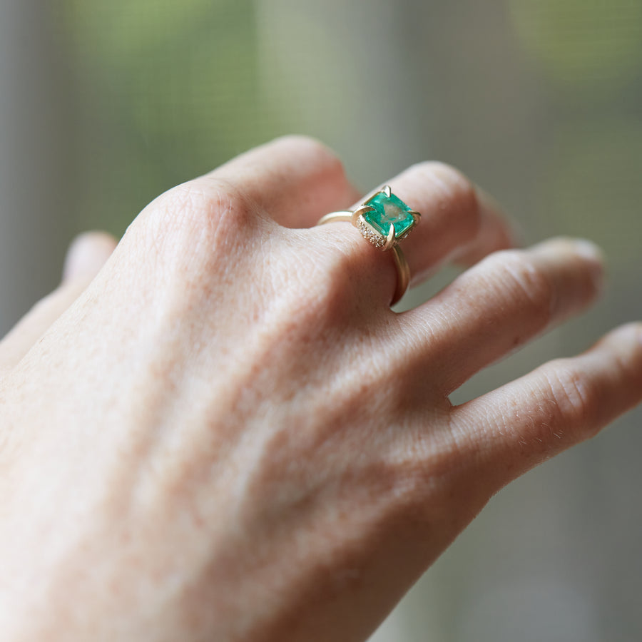 A 2.35ct Emerald ring with an 18k gold setting and shank. Four gold prongs secure the Emerald to the setting. The setting is inlaid with .32ct diamonds. The ring is displayed on a hand.