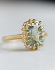 A 2.35ct Tourmaline ring set in 18k gold with a gold band. The Tourmaline is a stormy gray-blue. The Tourmaline is fastened to the setting by 16 gold prongs. The ring is viewed from the left side and is on a white background.