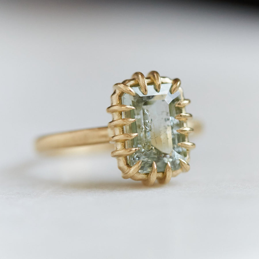 A 2.35ct Tourmaline ring set in 18k gold with a gold band. The Tourmaline is a stormy gray-blue. The Tourmaline is fastened to the setting by 16 gold prongs. The ring is viewed from the left side and is on a white background.