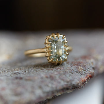 A 2.35ct Tourmaline ring set in 18k gold with a gold band. The Tourmaline is a stormy gray-blue. The Tourmaline is fastened to the setting by 16 gold prongs. The ring is viewed at an angle and is resting on a stone.