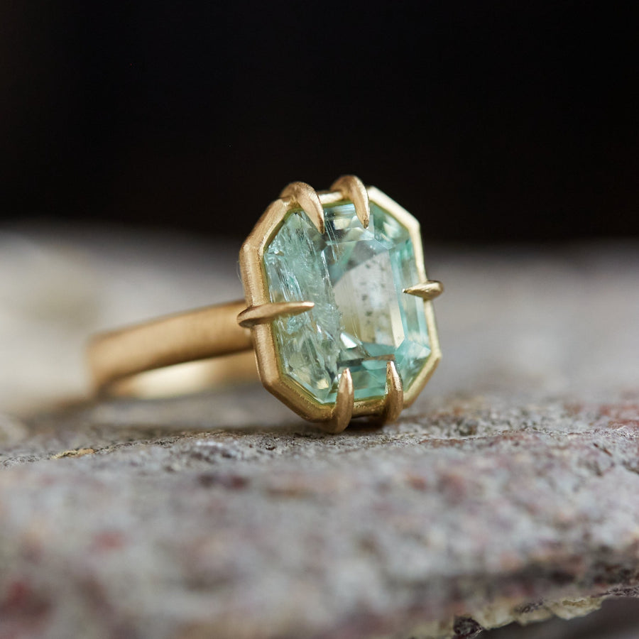 A 3.73ct Emerald ring. The emerald is a pale green and is set in 18k gold with an 18k gold band. The emerald is fastened to the setting by 6 prongs. The ring is viewed off center and displayed on a stone. 