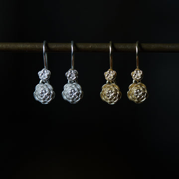 two pairs of drop earrings hanging beneath a black bar. The pair on the left are silver and the pair on the right is gold. The earrings are each of two roses with the top one smaller than the bottom.