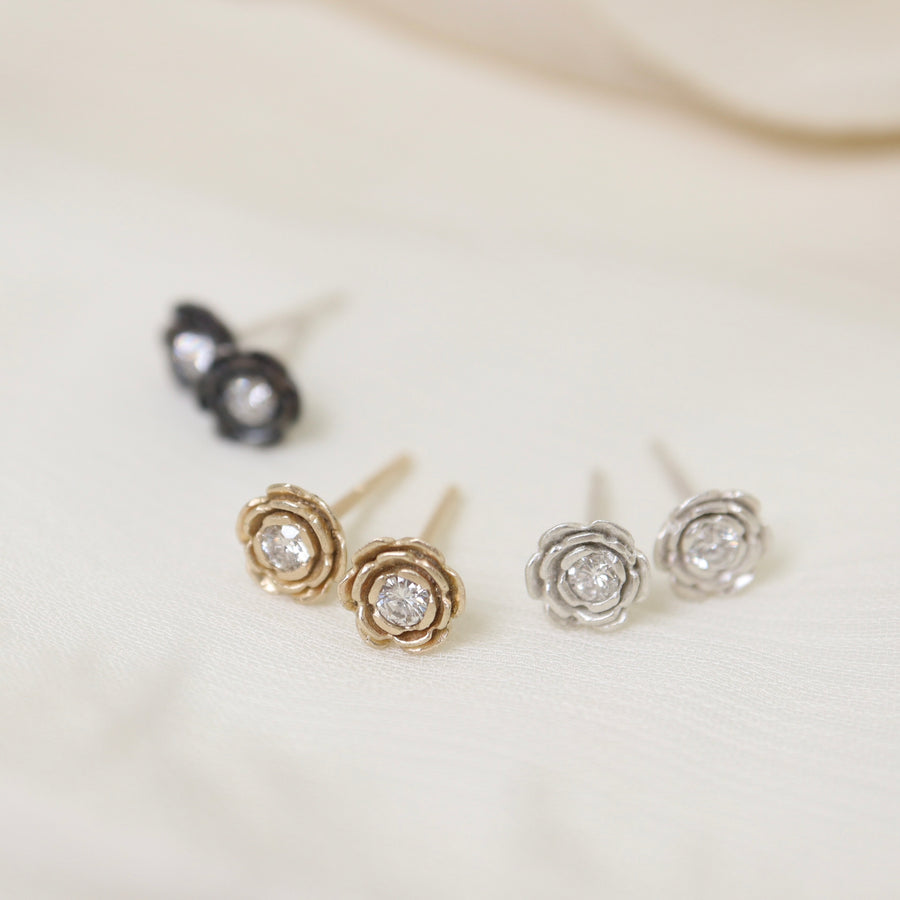 floral earrings studs made from metals of silver, gold, and oxidized silver are layed out on silk fabric. In the center of each floral stud is a diamond.