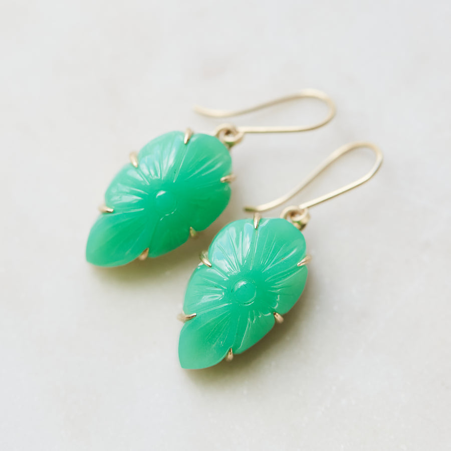 Two 17.93ct Carved Chrysoprase Earrings on a pale stone background. The carved Chrysoprase is set with six 14k gold prongs and gold ear wires. The Chrysoprase is shaped like a shield and carved with a flower motif.