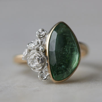 abstract green tourmaline rose cut gemstone set in a gold bezel with a bouquet of silver roses on one side. 