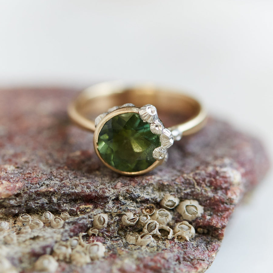 A 2.74ct tourmaline ring. The tourmaline is a deep green and is set in 14k gold with a 14k gold band. The rsetting is encrusted with bright sterling silver barnacles. The ring is viewed from the front and sits on a stone.