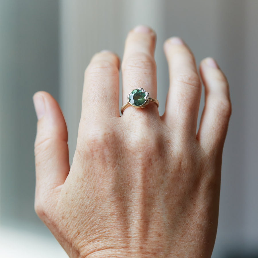 A 2.74ct tourmaline ring. The tourmaline is a deep green and is set in 14k gold with a 14k gold band. The rsetting is encrusted with bright sterling silver barnacles. The ring is displayed on a hand.