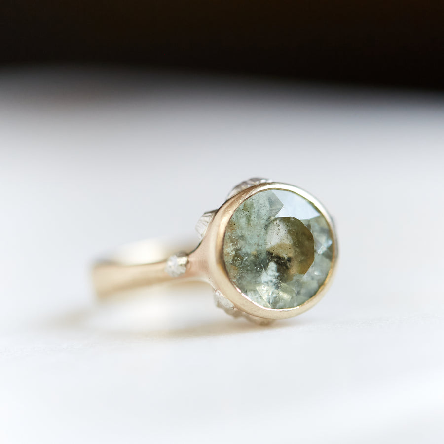 A 2.56ct Aquamarine ring. The ring is set in 14k gold with a tapered 14k gold band. The Aquamarine is a pale green with speckles. The setting is encrusted with bright sterling silver barnacles. The ring is on a white background. 