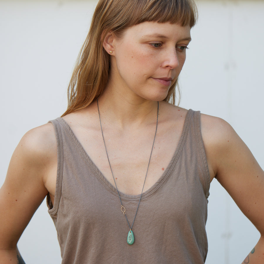 green turquoise teardrop bead with gold link and black chain. a scrimshaw hand is inked to the surface.