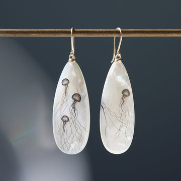 long teadrop mother of pearl earrings have gold ear wires and 3 jellyfish hand etched into the surface in black ink. A 4th jellyfish is on the back of the left earring, with the tentacles reach over onto the front.