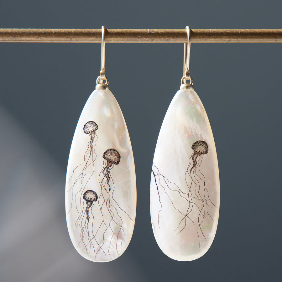 long teadrop mother of pearl earrings have gold ear wires and 3 jellyfish hand etched into the surface in black ink. A 4th jellyfish is on the back of the left earring, with the tentacles reach over onto the front.