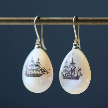 teardrop mother of pearl beads hang beneath gold ear wires. two different old whaling ships are hand etched into the surface with black ink via a method called scrimshaw.