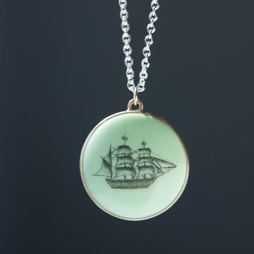 melon green round variscite cabochon is set in a gold bezel and hangs beneath a silver chain. an old whaling ship is hand-etched into the surface with black ink.