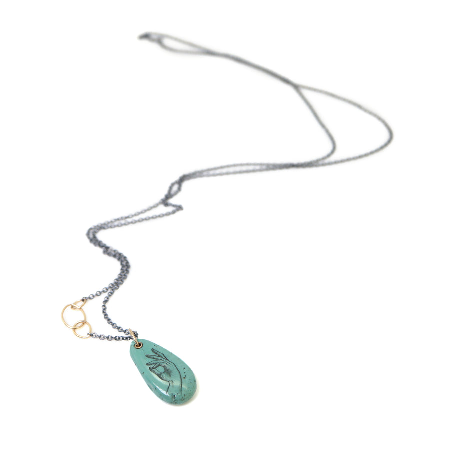 green turquoise teardrop bead with gold link and black chain. a scrimshaw hand is inked to the surface.