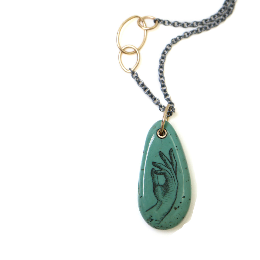 green turquoise teardrop bead with gold link and black chain. a scrimshaw hand is inked to the surface.