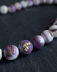 purple and white swirly beads of round quahog shells are strung on black silk and have gold barnacles attached throughout. A gold fish hook clasp attached the strand. Detail shot on black slate.