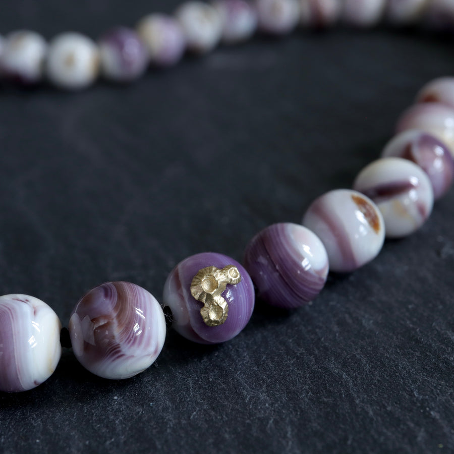 purple and white swirly beads of round quahog shells are strung on black silk and have gold barnacles attached throughout. A gold fish hook clasp attached the strand. Detail shot on black slate.