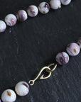 purple and white swirly beads of round quahog shells are strung on black silk and have gold barnacles attached throughout. A gold fish hook clasp attached the strand. Detail shot on black slate.