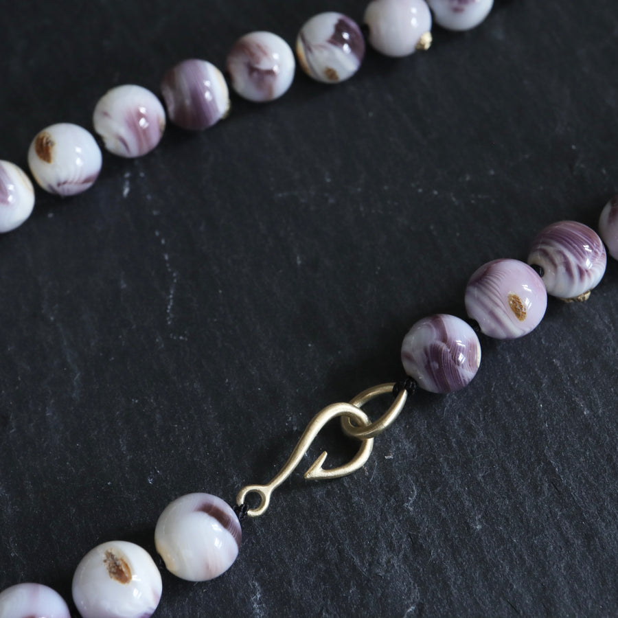 purple and white swirly beads of round quahog shells are strung on black silk and have gold barnacles attached throughout. A gold fish hook clasp attached the strand. Detail shot on black slate.